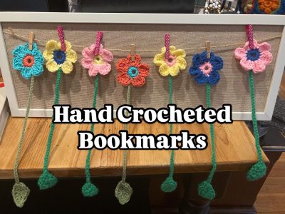 Six colorful hand-crocheted flower bookmarks hang on a display frame with clothespins on a wooden table.