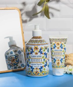 Hand wash and hand cream with decorative labels placed beside a mirror and seashell on a white surface.