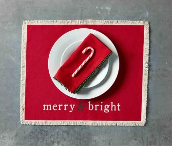 White plates with a red napkin and candy cane on a red placemat that reads "merry & bright" on a gray surface.