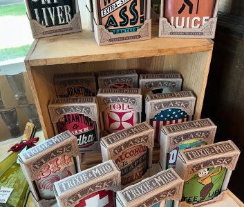 Wooden shelf displaying assorted Trixie & Milo flasks with various colorful and humorous designs and sayings.