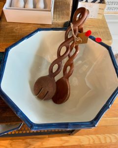 Wooden salad servers with twisted handles rest inside a white, blue-rimmed octagonal bowl on a wooden surface.