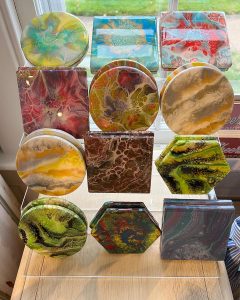 Display of colorful, abstract-patterned coasters in various shapes arranged on a transparent rack near a window.