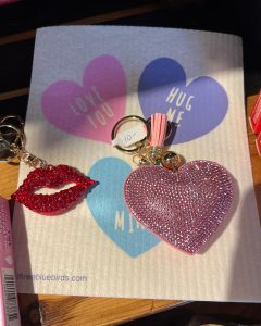 Keychains shaped like red lips and a pink heart displayed on a card with pastel hearts and romantic messages.