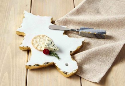 A cheese board with a cracker, blue cheese, a cherry tomato, and dill, next to a spreader and beige napkin on a wooden table.