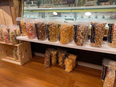 Bags of assorted flavored popcorn are displayed on white shelves in a shop with wooden floors.