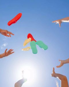 Four pairs of hands toss colorful flip-flops into a clear blue sky with sunlight shining from below.