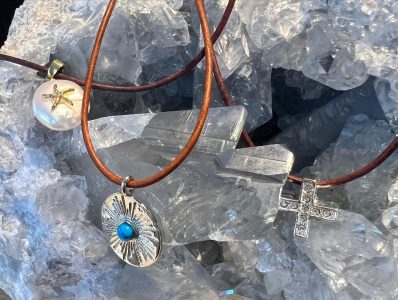 Three leather cord necklaces with pendants—a starfish, a turquoise disc, and a jeweled cross—on clear quartz crystals.