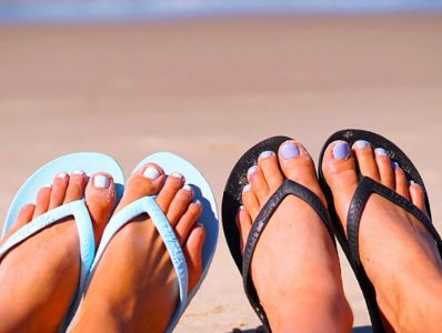 Two pairs of feet in flip-flops, one blue and one black, resting on sandy beach with blurred ocean in the background.