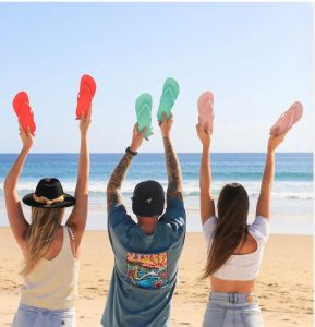 Three people on a beach hold up pairs of colorful flip-flops with the ocean and sky in the background.