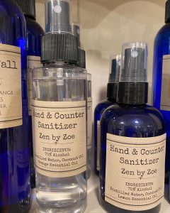 Bottles labeled "Hand & Counter Sanitizer Zen by Zoe" with ingredients listed, displayed on a shelf.