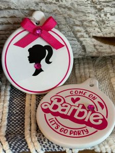 Two round ornaments featuring Barbie-themed designs with pink accents and rhinestones, placed on a woven surface.