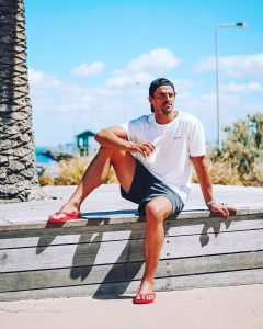 Man in a white t-shirt, shorts, and flip-flops sits on a wooden ledge outdoors on a sunny day.