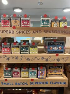Wooden shelves display various Spicewalla spice tins and collections, labeled "Superb Spices and Seasonings.