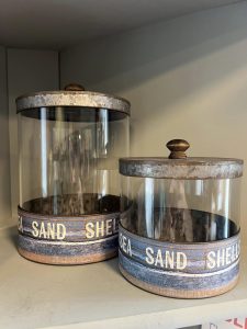Two empty glass jars with metal lids labeled "SEA SAND SHELLS" sit on a shelf.