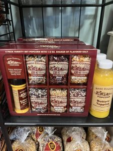 A display of Amish Country popcorn kits with assorted popcorn, flavored salts, and butter topping bottles on a shelf.