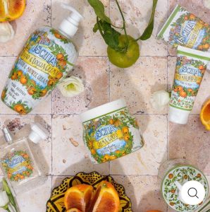 Skincare products with colorful labels are arranged on a tiled surface with citrus fruits, a flower, and a patterned dish.