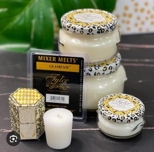 Four GlamFam scented candles and one wax melt pack are arranged on a table with a green leaf in the background.