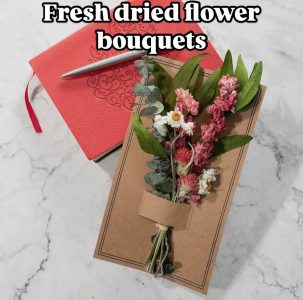 A small bouquet of dried flowers on a brown box, beside a red notebook and pen on a white marble surface.