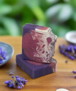 Two artisanal soap bars with swirled purple, red, and white patterns are stacked on a wooden surface with lavender nearby.