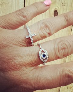 A hand with pink nail polish wearing a cross-shaped ring and an eye-shaped ring with a dark center stone.