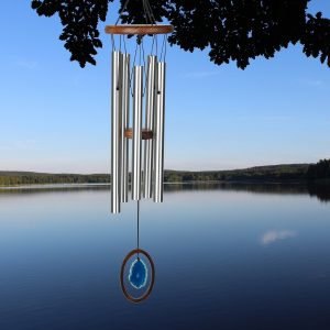 Silver wind chime with a blue stone pendant hangs in front of a calm lake and forested shoreline under a clear sky.