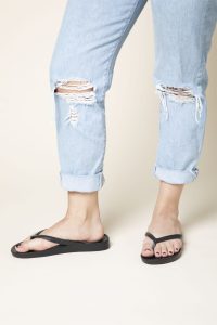 Person wearing light blue ripped jeans and black flip-flops standing on a light-colored surface.