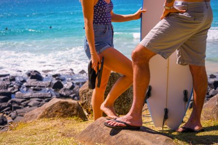 Two people in summer clothes stand on rocks by the ocean, one holding a surfboard and the other holding flip-flops.