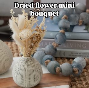 A small white vase holds a dried flower arrangement on a table, with blue decorative beads and books in the background.