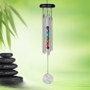 A wind chime with metal tubes and colorful beads hangs over water, with bamboo leaves and stacked stones in the background.