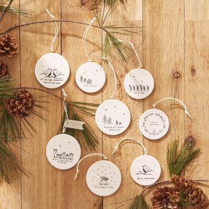 Nine round white ceramic Christmas ornaments with black illustrations and text are arranged on a wooden surface with pine branches.