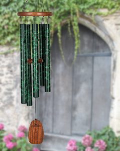 Green and black wind chimes with leaf patterns hang in front of a weathered wooden door and blooming pink flowers.
