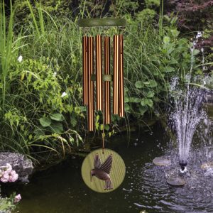 A wind chime with a hummingbird design hangs above a garden pond with a small fountain and green plants.