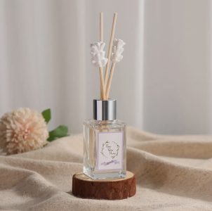 A glass reed diffuser with white floral sticks sits on a wooden base, with a flower and fabric in the background.