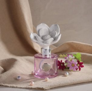 A pink perfume bottle with a white flower-shaped cap sits on beige fabric, surrounded by artificial flowers and pearls.