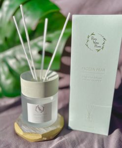 A glass reed diffuser labeled "Frozen Pear" sits next to its packaging on a wooden hexagonal coaster.