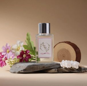 A perfume bottle labeled "Pretty Valley Home" is displayed on a rock with flowers, greenery, and a wood slice.