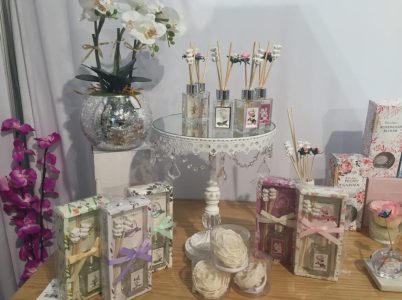 A display of reed diffusers, decorative boxes, artificial flowers, and roses on a wooden table with a white backdrop.