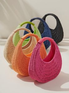 Five woven handbags in pink, orange, natural, green, and navy are arranged upright on a light-colored surface.