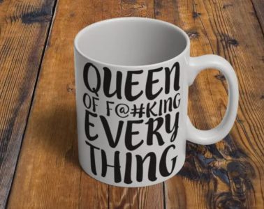 A white mug on a wooden table with the text "Queen of F@#king Everything" printed in bold black letters.