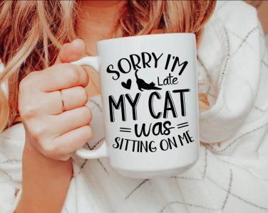 A person holds a mug that reads, "Sorry I'm late, my cat was sitting on me," with a heart and cat graphic.