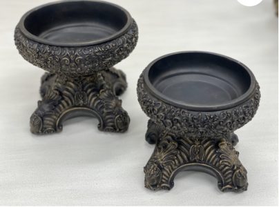 Two ornate, round black bowls with intricate gold detailing on decorative pedestal bases, placed on a light surface.