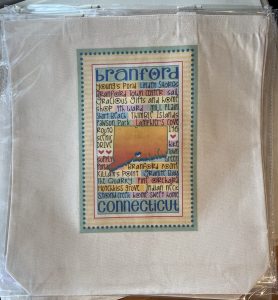 A beige tote bag features a colorful graphic listing Branford, Connecticut landmarks and attractions.