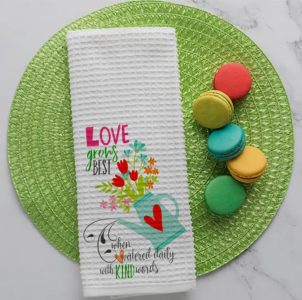A white towel with a watering can and flower design, placed on a green mat next to colorful macarons.