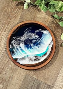 Round wooden tray with resin art depicting ocean waves, placed on a wood floor next to green leafy plants.
