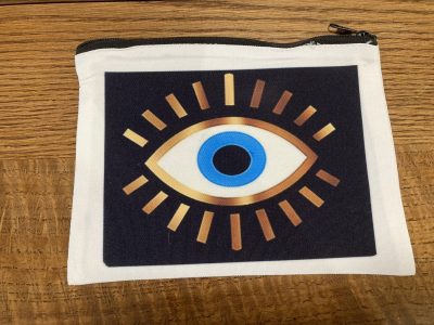 A white zippered pouch featuring a stylized eye design with gold rays and a blue center, placed on a wooden surface.