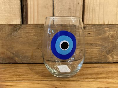 A clear glass with a blue evil eye design sits on a wooden surface against a wood panel background.