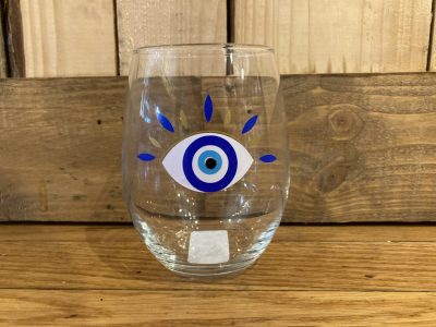 A clear glass with a blue and white eye design and blue and gold lines, placed on a wooden surface in front of a wood wall.