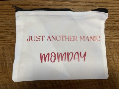 A white zippered pouch with the text "JUST ANOTHER MANIC MOMDAY" in pink lettering on a wood surface.