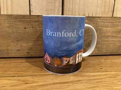 A coffee mug with an illustrated town scene and the text "Branford, CT" sits on a wooden surface.