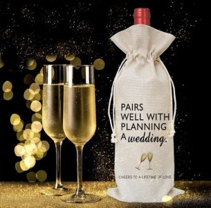 Two champagne glasses and a wine bottle in a cloth bag labeled "Pairs well with planning a wedding" with gold lights in background.